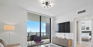 A modern hotel room with a large window, a flat-screen TV, a sofa, a chair, and a small table. - Bay-View High-Rise: Private Balcony, Pool & Gym (Miami)