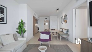 A modern living room with a sofa, a dining table, and a kitchen in the background. - Bay-View High-Rise: Private Balcony, Pool & Gym (Miami)