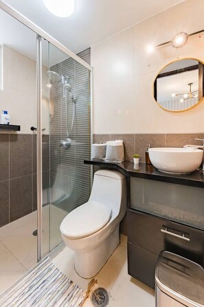 Une salle de bain moderne avec une douche à parois de verre, des toilettes blanches, un lavabo avec un miroir rond et des meubles aux tons foncés.