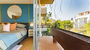 A bedroom with a bed, bedside table, and a large mirror. There is a balcony with a view of the city.