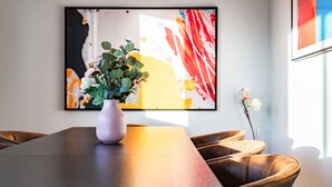 En spiseplads med et langt bord, en vase med blomster og et stort abstrakt billede på væggen.