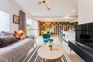 A modern living room with a sofa, a round coffee table, a TV, and a kitchen area with a bar and chairs. - Penthouse in Roma Norte with City Skyline View (Mexico City)