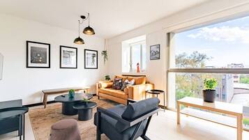 A modern living room with a sofa, coffee table, and chairs, a balcony with a view, and framed artwork on the walls.
