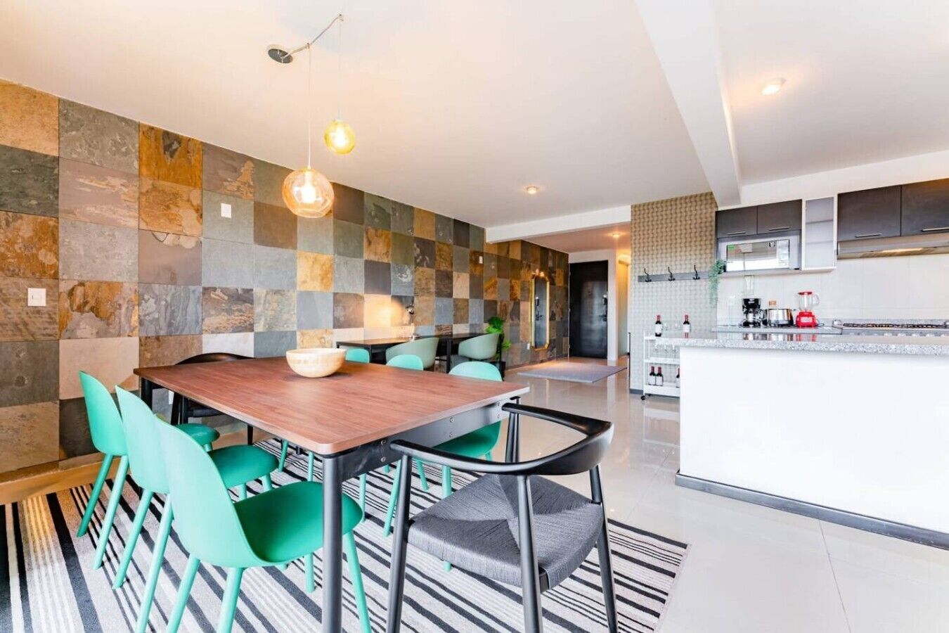 A modern kitchen with a central island, a dining area with a wooden table and green chairs, and a stone-patterned wall.