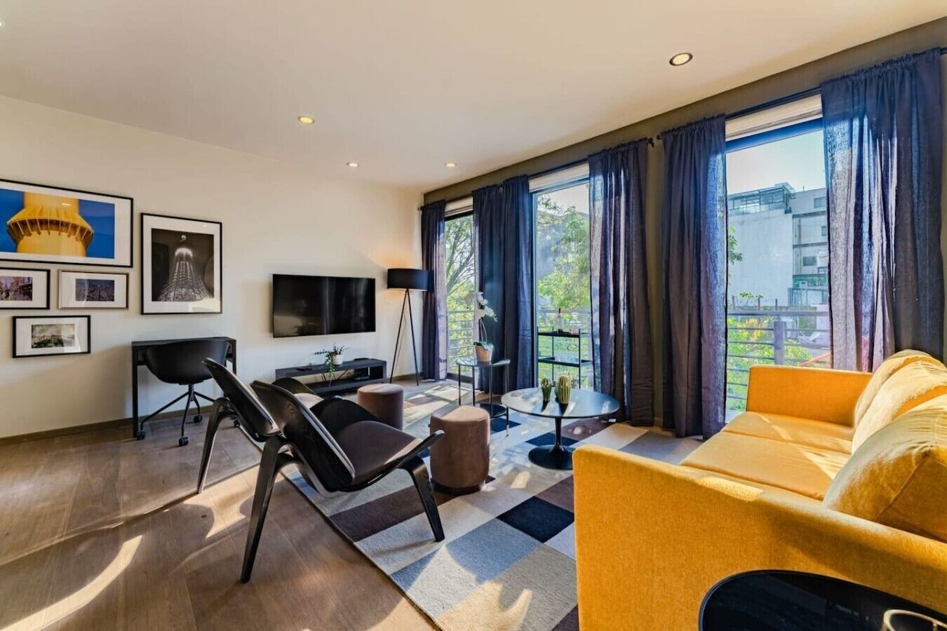 A modern living room with a sofa, a TV, a coffee table, and a chair. There is a window with curtains, a plant on a side table, and artwork on the wall.