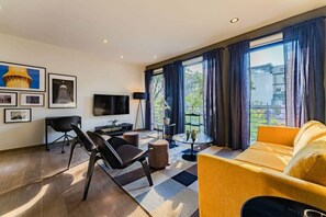 A modern living room with a sofa, a TV, a coffee table, and a chair. There is a window with curtains, a plant on a side table, and artwork on the wall. - Roma Norte Condo: Balcony, Parking & City Views (Mexico City)