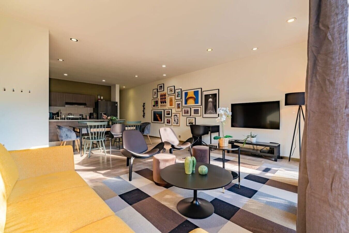 A modern living room with a sofa, chairs, a coffee table, a TV, and a kitchen in the background.