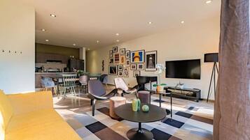 A modern living room with a sofa, chairs, a coffee table, a TV, and a kitchen in the background.