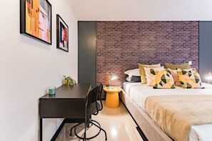 A modern bedroom with a brick accent wall, a bed with patterned pillows, a desk with a chair, and a small side table. - Roma Norte Condo with City Views & Workspace (Mexico City)