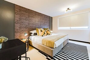 A modern bedroom with a brick accent wall, a large bed with patterned pillows, a desk with a lamp, and a black and white striped rug. - Roma Norte Condo with City Views & Workspace (Mexico City)