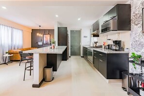 A modern kitchen with a central island, dark cabinetry, and a dining area with a sofa and chairs. - Roma Norte Condo with City Views & Workspace (Mexico City)