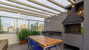 A rooftop patio with a wooden table and blue chairs, a grill, and a city view. - Studio in Historic Center Near Santa Casa Tco0712 (Porto Alegre)
