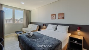 Ett hotellrum med en stor säng, ett litet sängbord, en stol och utsikt över staden genom stora fönster.