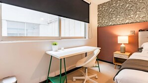 Ein Hotelzimmer mit Schreibtisch, Stuhl, Bett, Nachttisch mit Lampe und einem Fenster mit Jalousien.