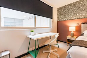 Ein Hotelzimmer mit Schreibtisch, Stuhl, Bett, Nachttisch mit Lampe und einem Fenster mit Jalousien.