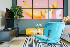 Ein modernes Wohnzimmer mit Fernseher, blauem Sessel, gelber Couch und einem gestreiften Teppich.