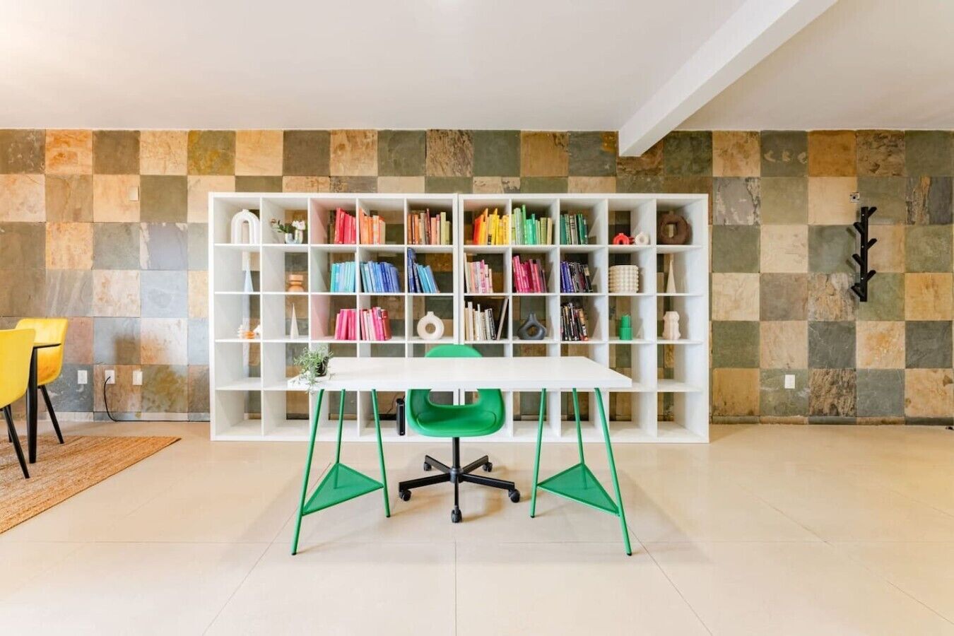 Una habitación con un escritorio blanco, sillas verdes, una estantería blanca y una pared con azulejos.