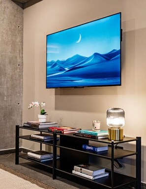 A modern living room with a large flat-screen TV mounted on the wall, a black entertainment unit with books and decorative items, and a small table with a lamp. - Stylish 1BR • Gym & Workspace • Near Reforma (Mexico City)