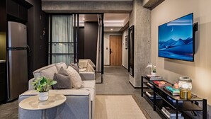 A modern living room with a sofa, a coffee table, and a TV. There is a bookshelf with books and a decorative lamp. The room has a minimalist design with a large window and a door leading to another room. - Stylish 1BR • Gym & Workspace • Near Reforma (Mexico City)