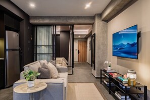 A modern living room with a sofa, a coffee table, and a TV. There is a bookshelf with books and a decorative lamp. The room has a minimalist design with a large window and a door leading to another room. - Stylish 1BR • Gym & Workspace • Near Reforma (Mexico City)