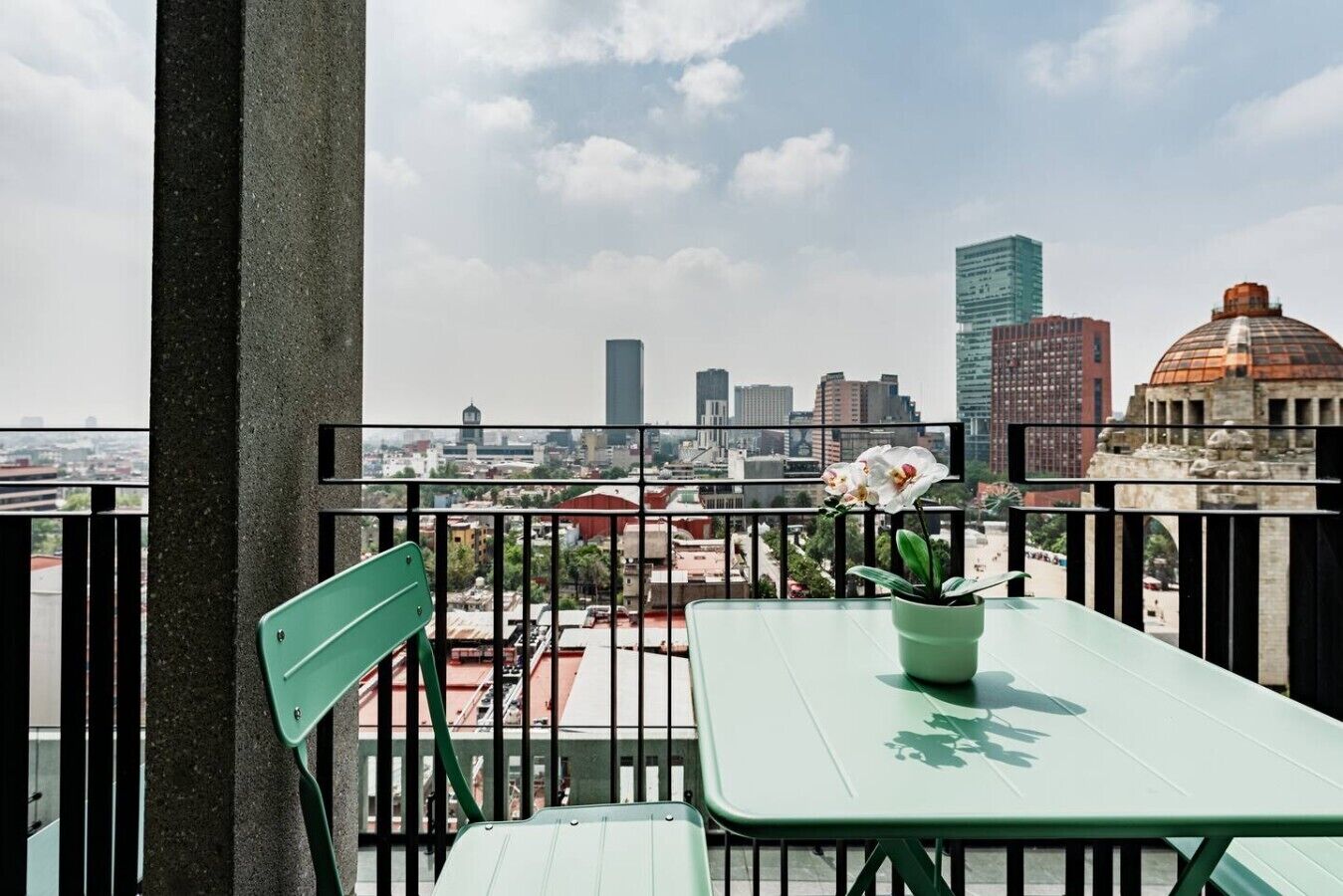 En balkon med et grønt bord og stol, en plantekasse og udsigt over bybilledet.