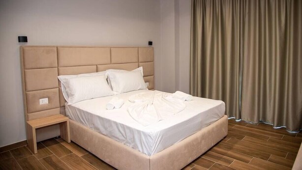 A neatly made bed with a headboard, a bedside table, and a curtain on the window. - Hotel Lubjana (Durrës)