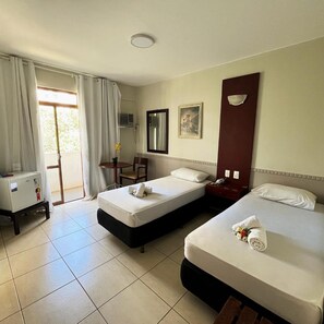 Quarto de hotel com duas camas, uma escrivaninha, uma cadeira, janela com cortinas e ar condicionado fixado na parede.
