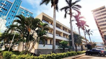 Um edifício de vários andares com varandas, rodeado por palmeiras e um espaço pavimentado com um carro estacionado.