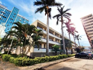 Un edificio de varios pisos con balcones, rodeado de palmeras y un área pavimentada con un coche estacionado.
