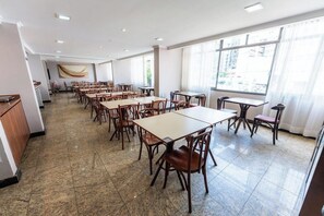 Un comedor amplio con varias mesas y sillas, ventanas grandes con cortinas y suelo de baldosas.