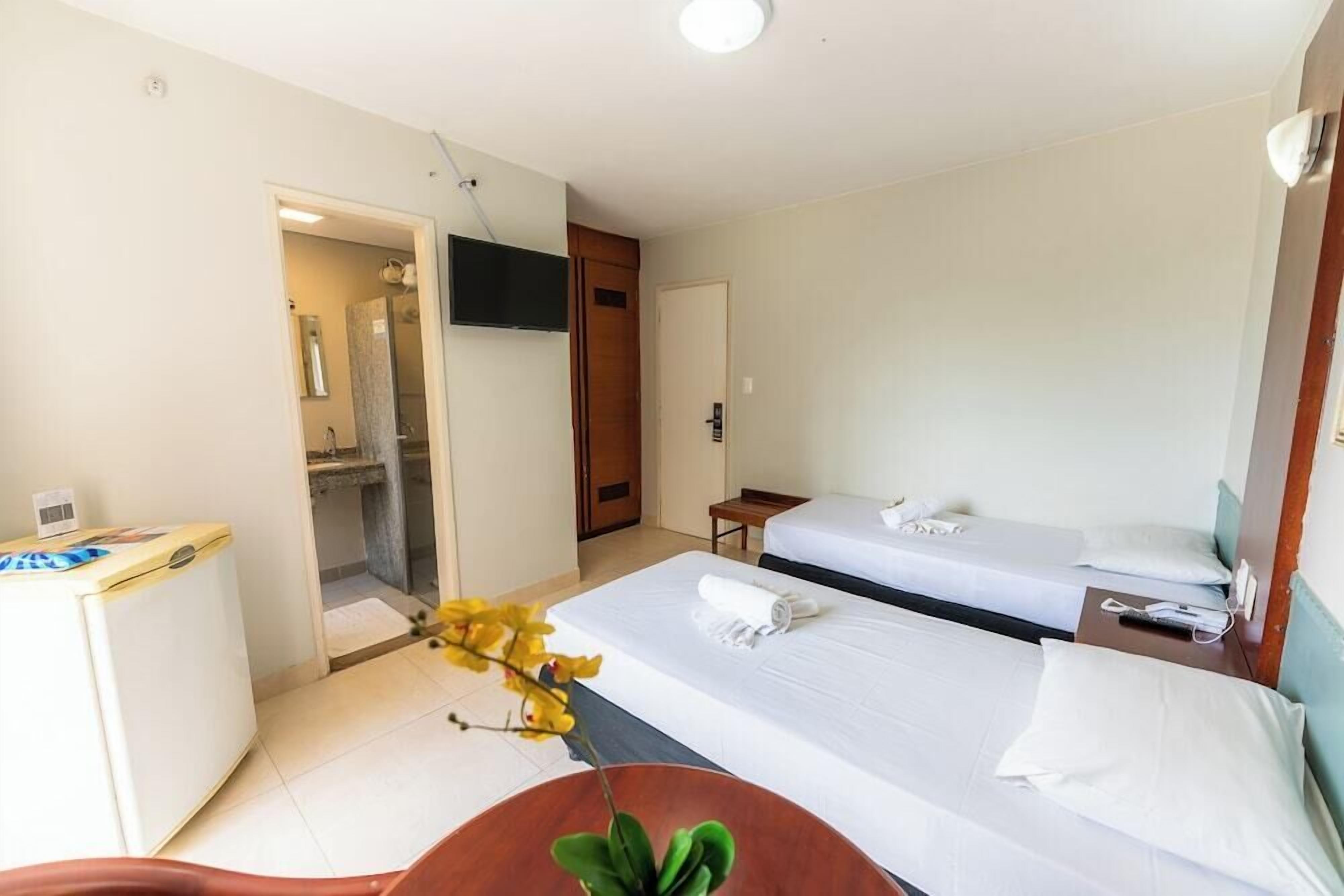 Quarto de hotel com duas camas, uma escrivaninha, televisão e banheiro visível por uma porta aberta.