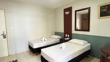 Quarto de hotel com duas camas de solteiro, uma escrivaninha, uma cadeira, um espelho e uma porta.