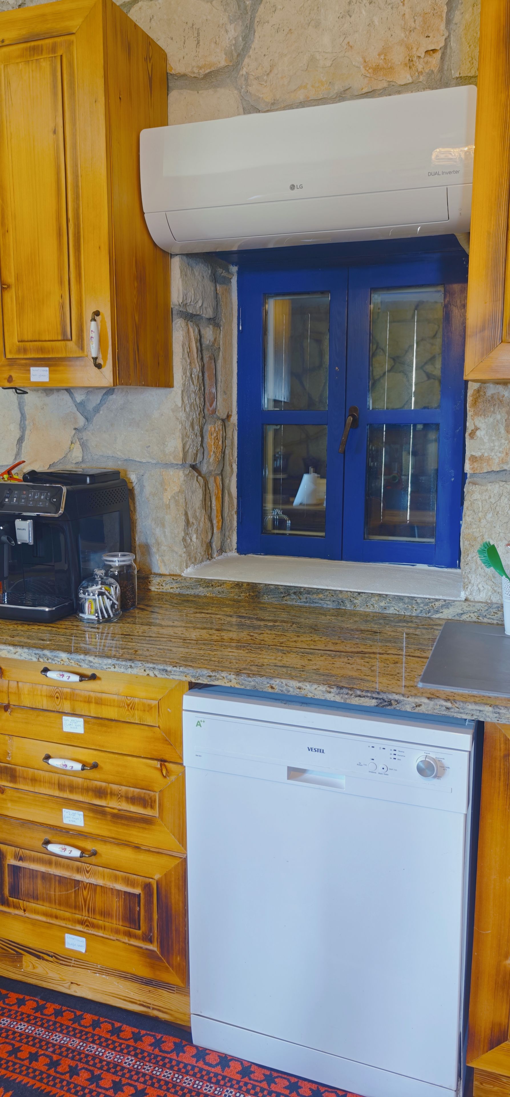 Cocina con pared de piedra, armarios de madera, puerta azul, lavavajillas blanco y cafetera.
