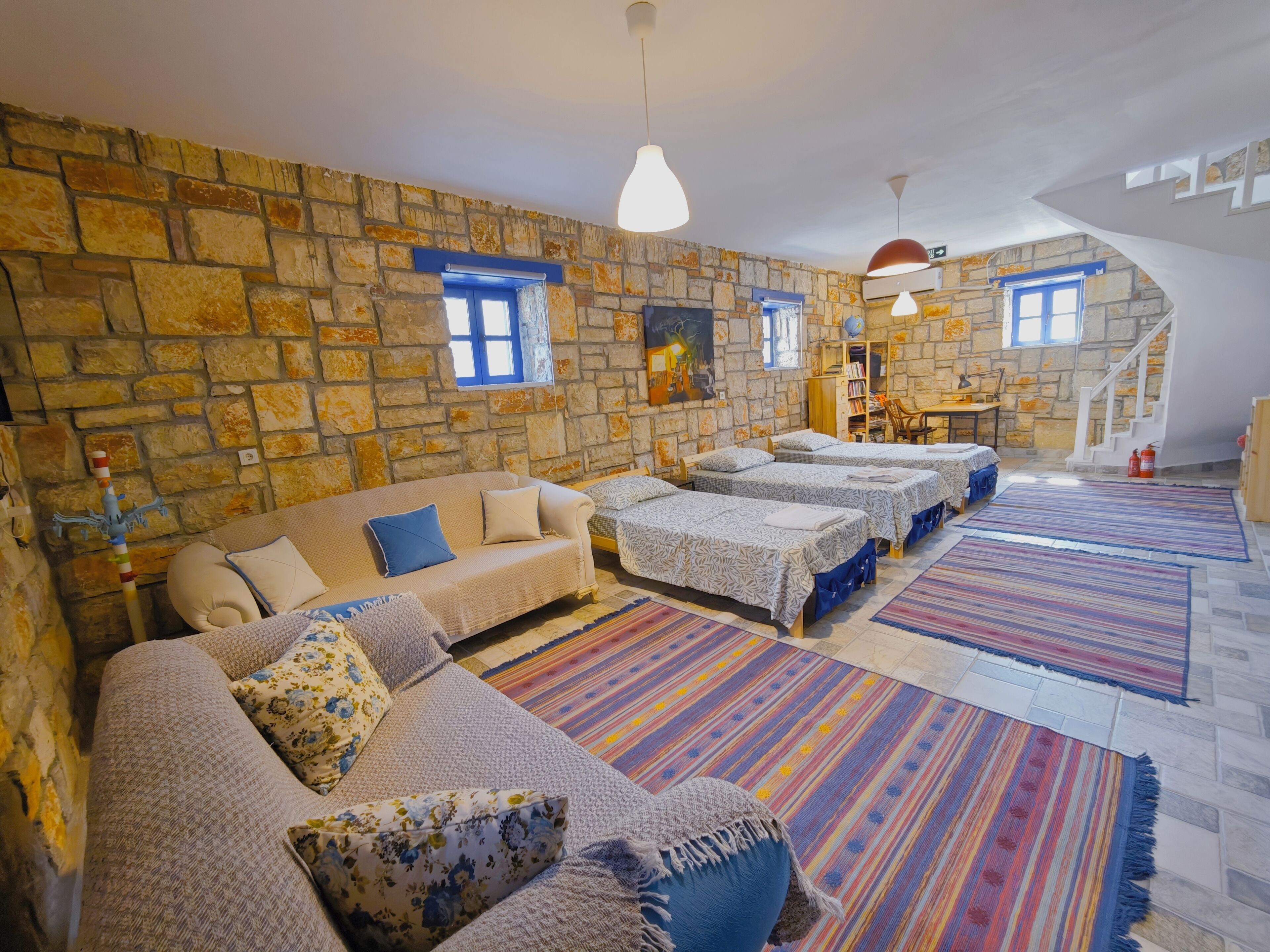 Una habitación acogedora con pared de piedra, un sofá, varias camas, una escalera y una alfombra colorida.