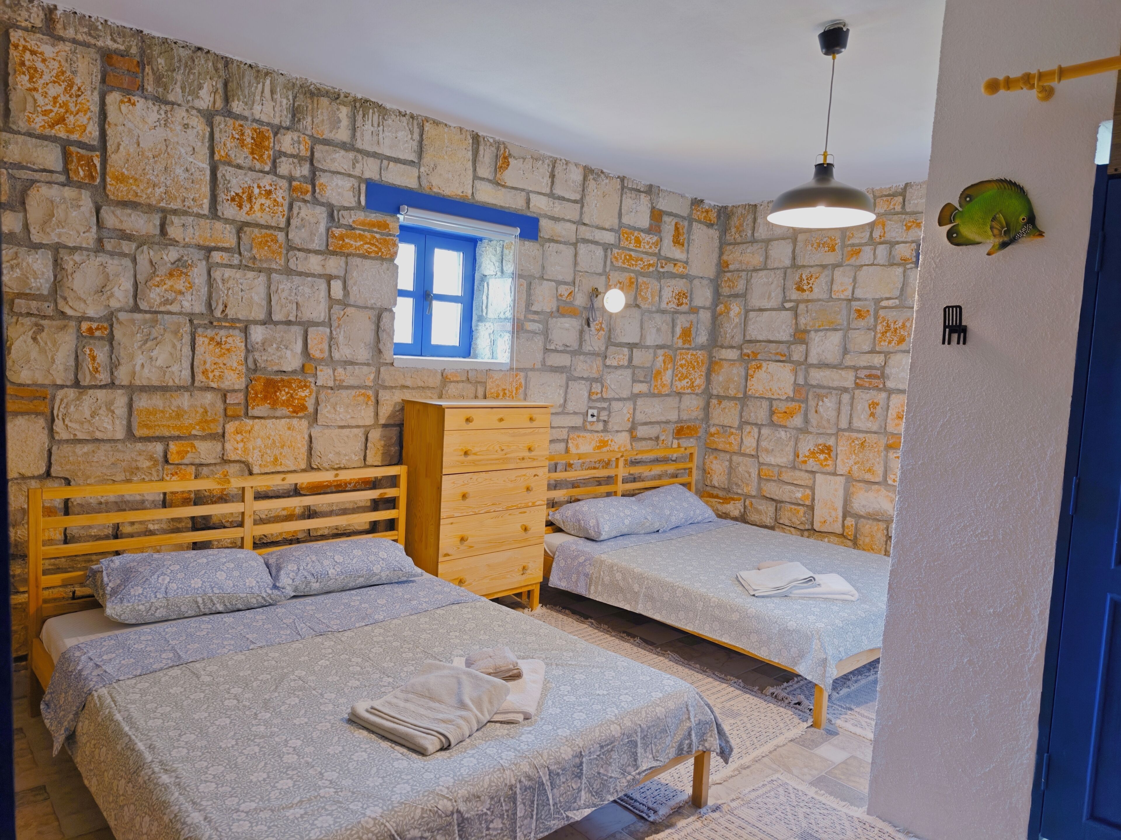 A room with two beds, a wooden dresser, a stone wall, a window with blue shutters, and a hanging light fixture.