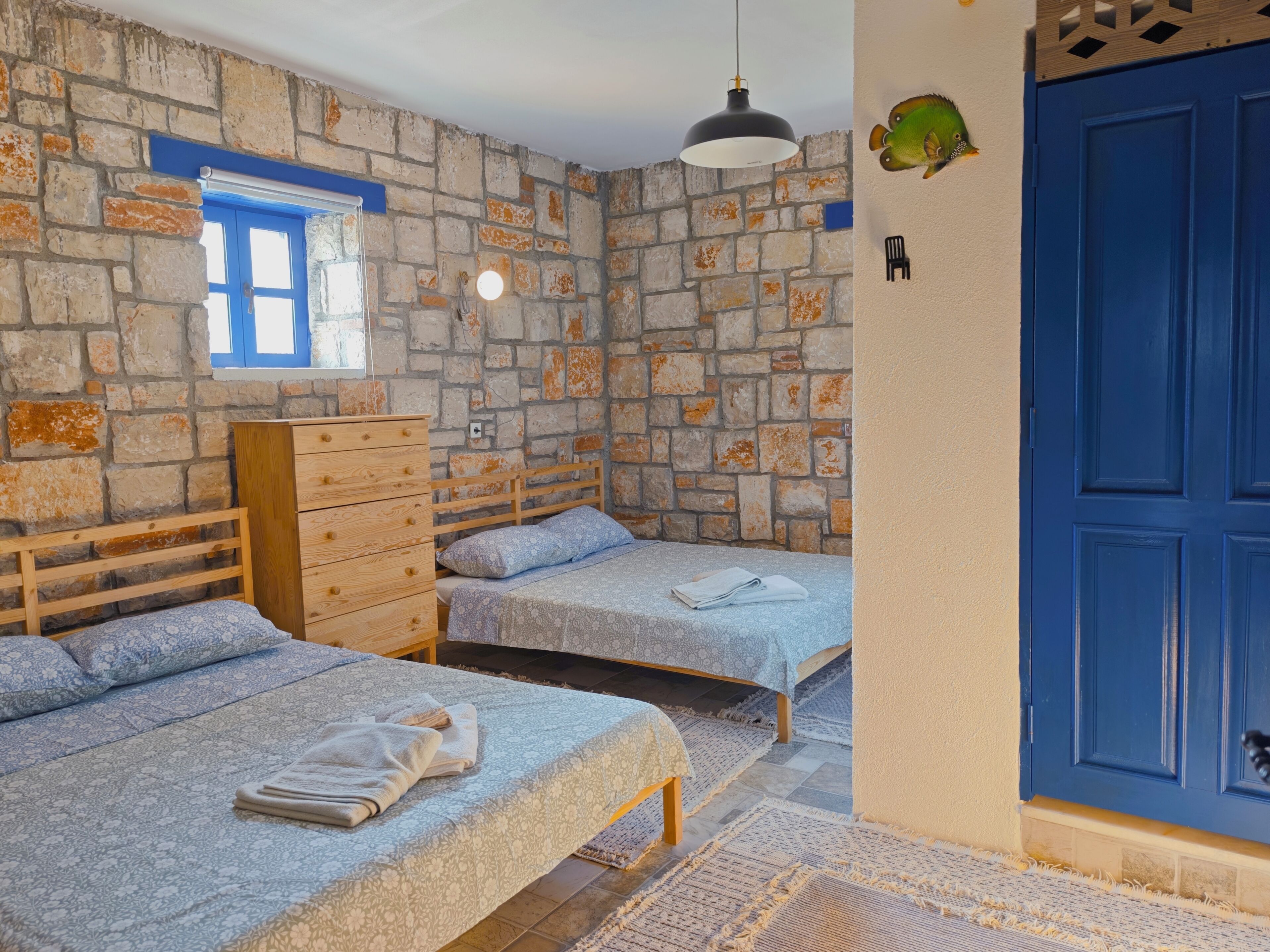 A room with two beds, a dresser, a blue door, and a stone wall.