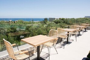 An outdoor terrace with tables and wicker chairs, overlooking a coastal landscape.