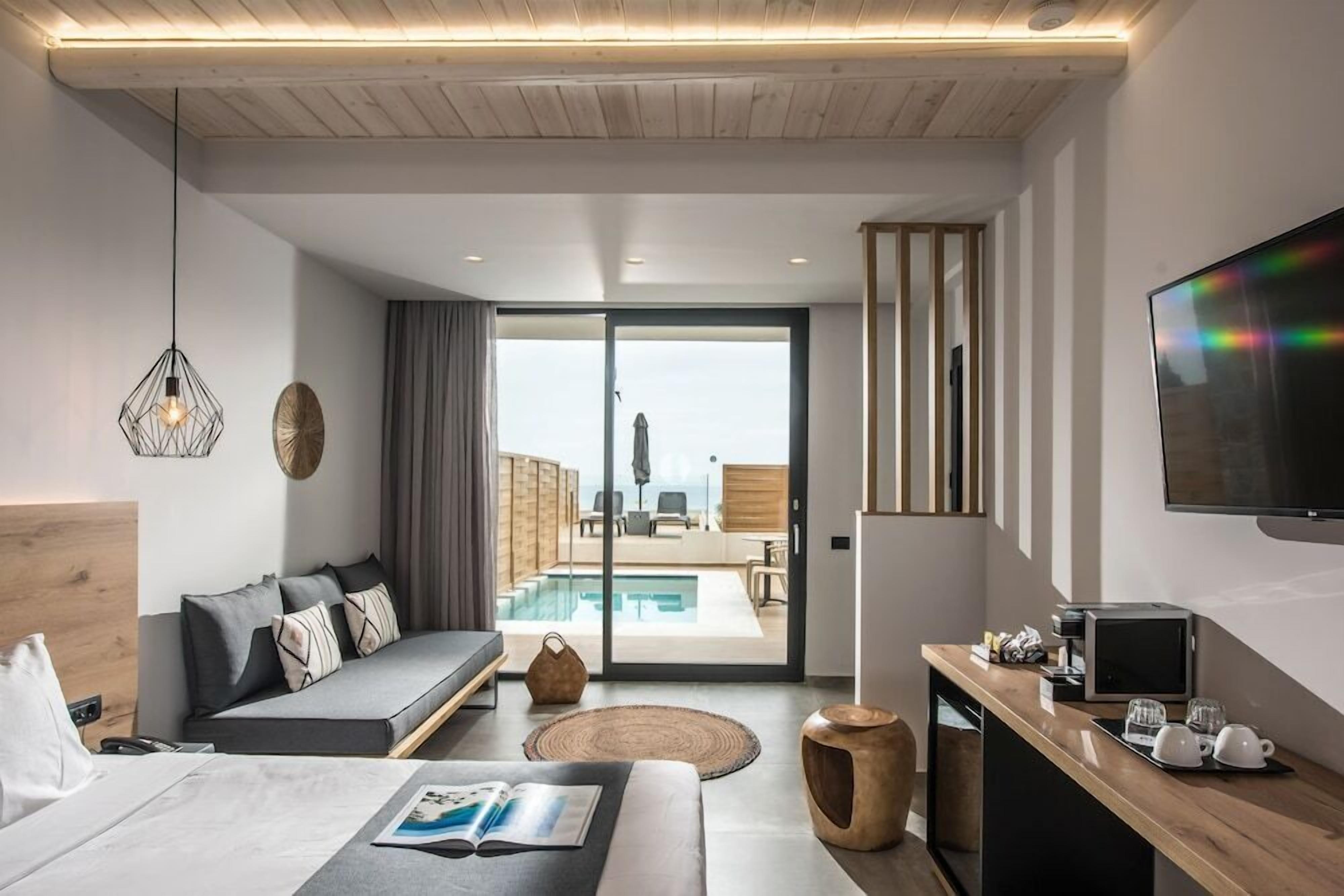 Een moderne hotelkamer met een bed, een sofa, een bureau met een computer, een televisie en uitzicht op een buitenzwembad.