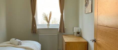 A hotel room with a bed, a bedside table, a radiator, a window with curtains, a wall-mounted picture, and a wooden wardrobe.