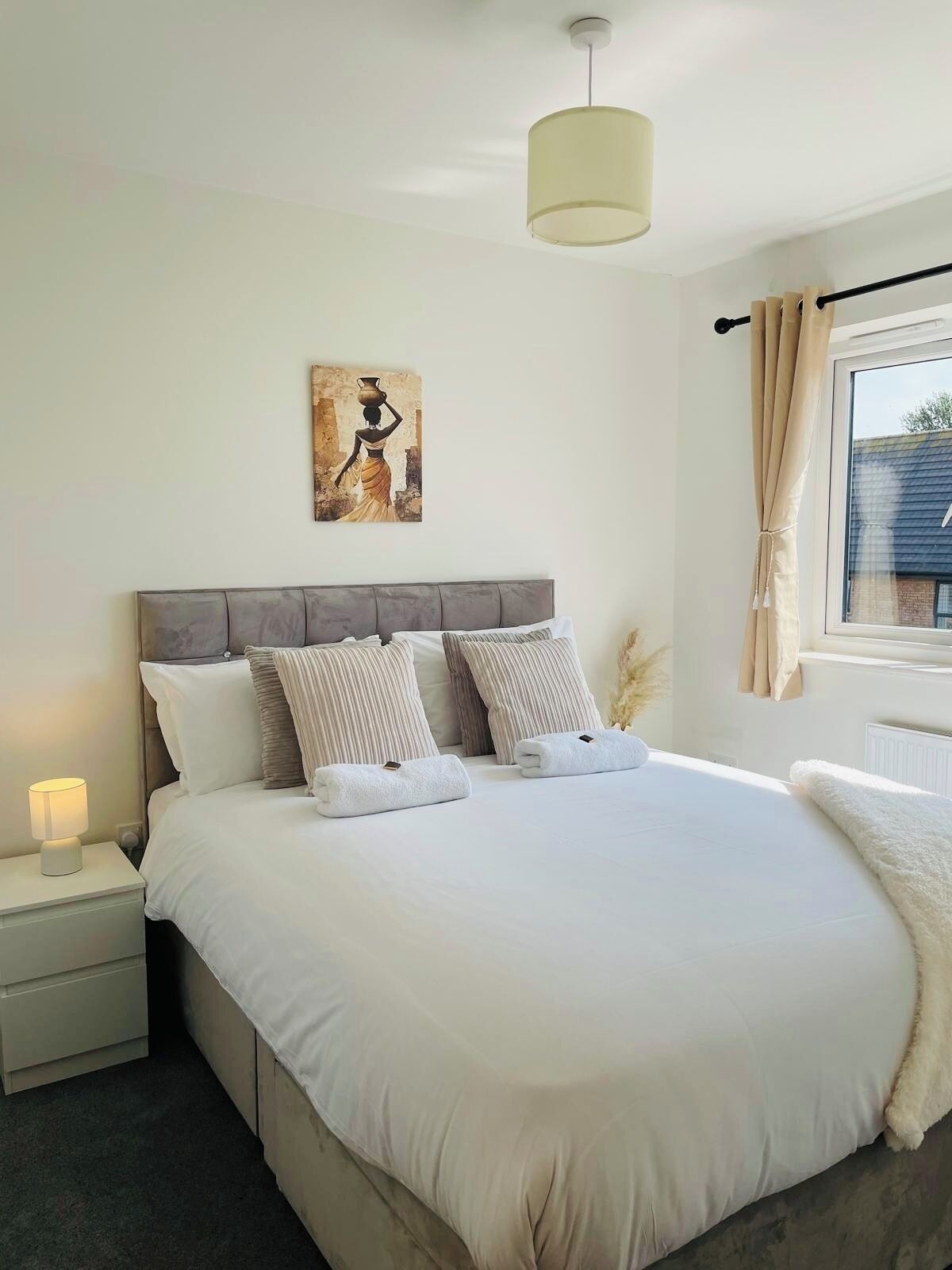 A neatly made bed with pillows and a bedside lamp, a painting on the wall, and a window with curtains.