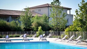 Piscina com espreguiçadeiras, um edifício com telhado em azulejos e vegetação ao redor.