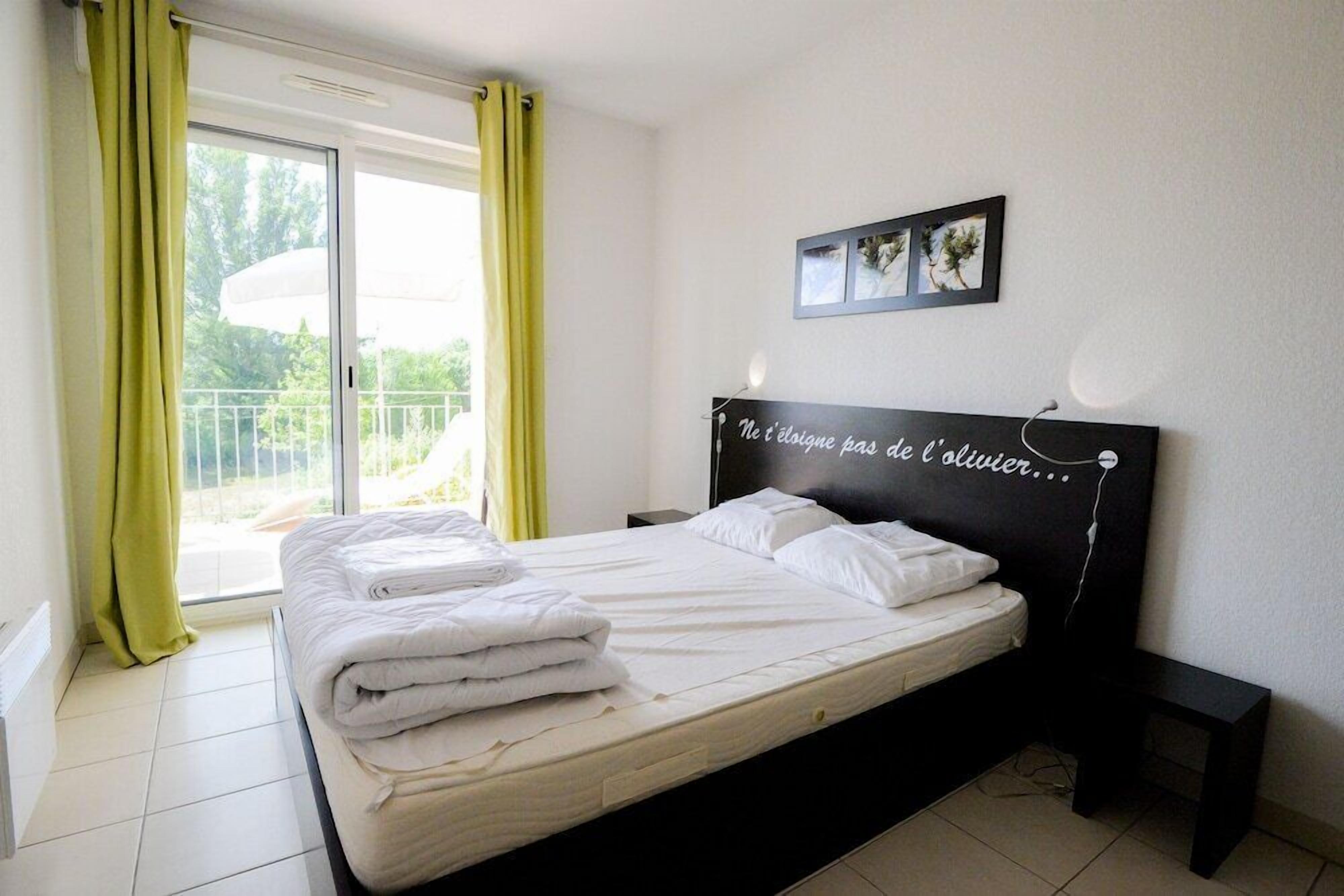 Une chambre à coucher avec un lit, une table de chevet, une lampe fixée au mur et une fenêtre avec des rideaux.