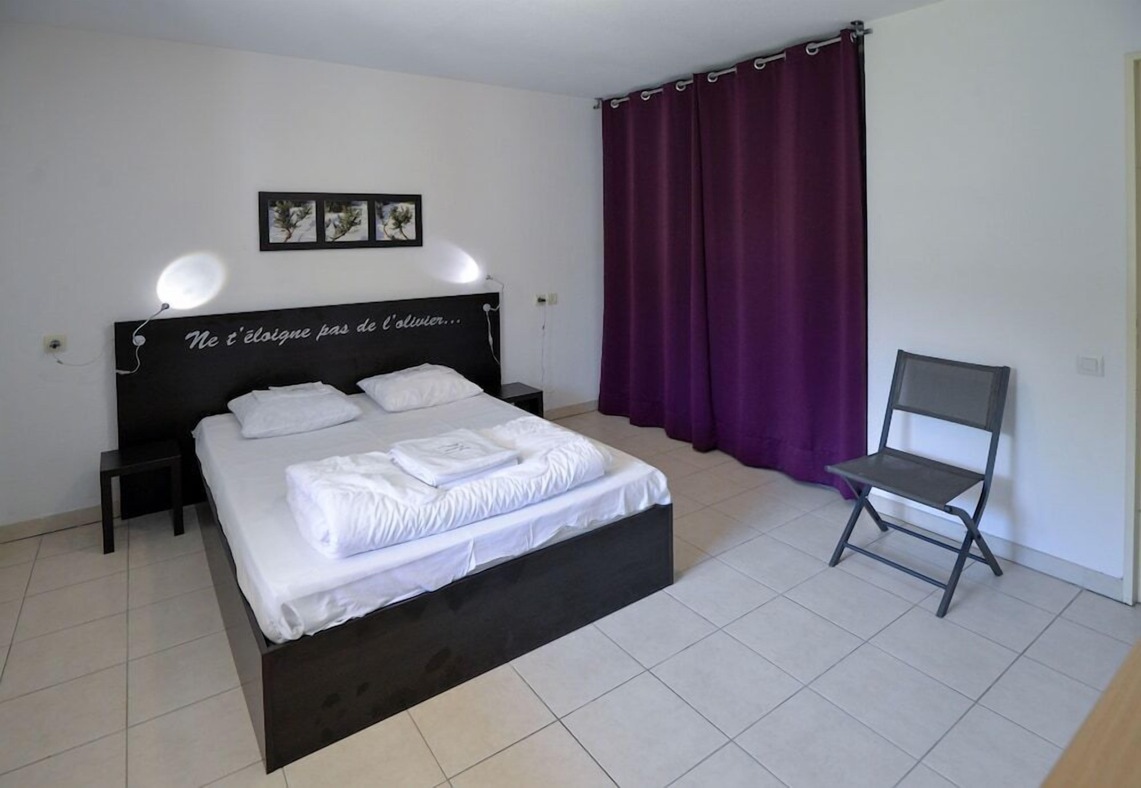 Une chambre à coucher comprenant un lit, une table de chevet, une chaise, une lampe fixée au mur et un rideau.