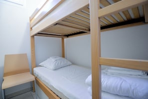 Una cama litera con estructura de madera, una cama individual con ropa de cama blanca y una silla con cojín de color claro.