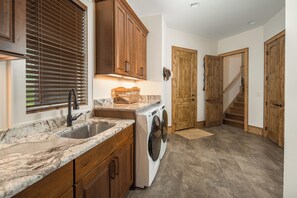 A kitchen with a sink, countertop, wooden cabinets, a washing machine, and a door leading to a staircase. - Buffalo Whiskey Soak Above the Clouds at a Luxe 5BR Eagle's Nest Lodge (Banner Elk)
