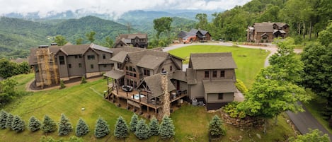 A large, multi-story building with a spacious deck and outdoor hot tub, surrounded by lush greenery and mountains in the background.