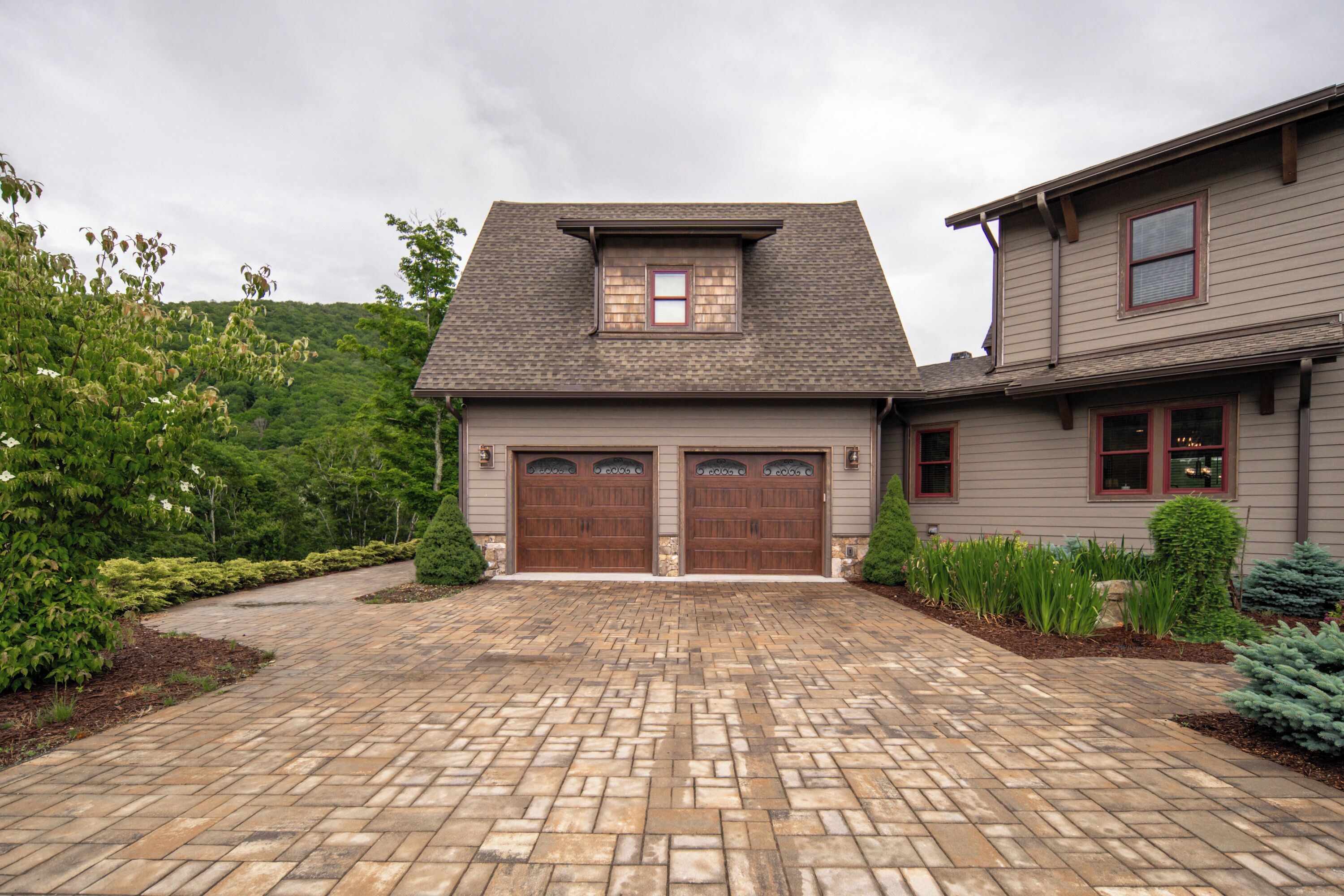 A two-story house with a double garage, a stone pathway leading to the entrance, and a landscaped garden with various shrubs and a small tree.