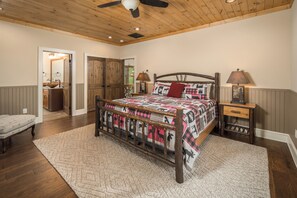 A bedroom with a wooden bed frame, a quilt with animal prints, a wooden nightstand, and a ceiling fan. - Buffalo Whiskey Soak Above the Clouds at a Luxe 5BR Eagle's Nest Lodge (Banner Elk)