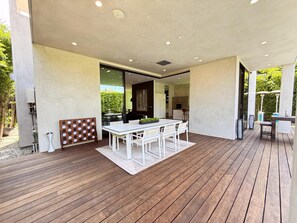 An open-plan living area with a wooden deck, a dining table with chairs, and a kitchen visible through an open doorway. - Modern Museum 5-bedroom infinity pool (Los Angeles)