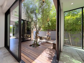 A modern patio with wooden flooring, a small tree, wicker furniture, and a view of a lush garden through large windows. - Modern Museum 5-bedroom infinity pool (Los Angeles)